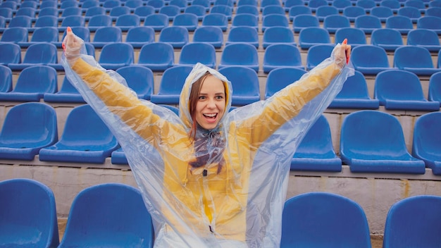 Junge Frau im Regenmantel, die allein auf der Stadiontribüne sitzt und für die Lieblingssportmannschaft jubelt, die sich freut, bei Regenwetter zu gewinnen