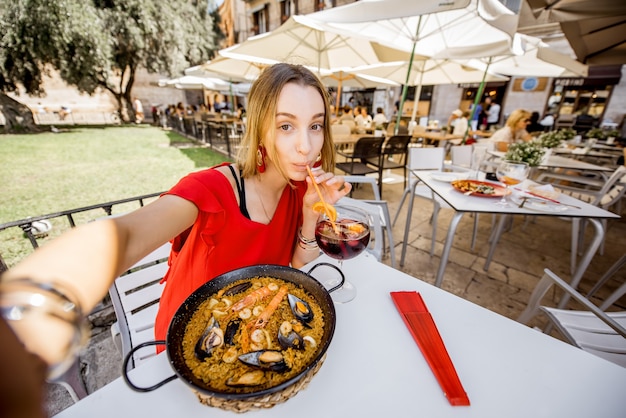 Junge Frau im roten Kleid, die ein Selfie-Foto mit Meer-Paella, traditionellem valencianischen Reisgericht und Sangria-Getränk macht, das draußen im Restaurant in Valencia sitzt?