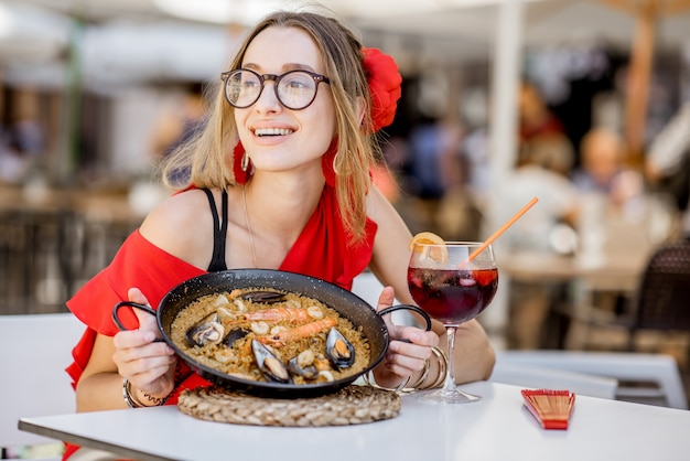 Junge Frau im roten Kleid isst Meer-Paella, traditionelles valencianisches Reisgericht, sitzt draußen im Restaurant in Valencia, Spanien