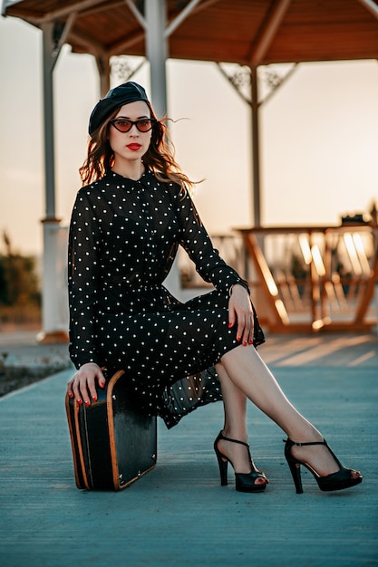 Junge Frau im schwarzen Tupfenkleid der Weinlese mit dem Retro- Koffer in der Hand, der draußen aufwirft