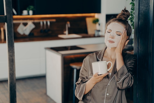 Junge Frau im Seidenpyjama, die zu Hause bleibt und morgens eine Tasse Kaffee genießt