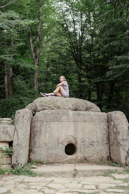 Junge Frau im Sommerkleid sitzt auf einem großen Dolmenstein im Wald und schaut weg