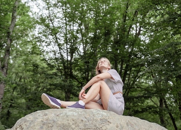 Junge Frau im Sommerkleid sitzt auf einem großen Stein im Wald und schaut weg