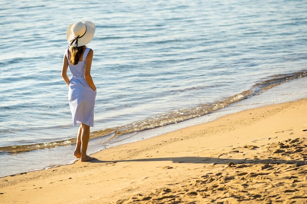 Junge Frau im Strohhut und in einem Kleid, das allein auf leerem Sandstrand am Seeufer geht