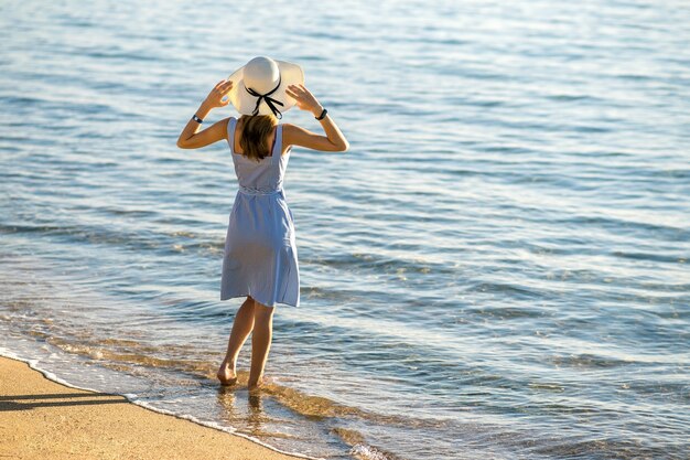 Junge Frau im Strohhut und in einem Kleid, das allein auf leerem Sandstrand am Seeufer steht. Einsames Touristenmädchen, das Horizont über ruhiger Ozeanoberfläche auf Urlaubsreise betrachtet.