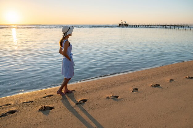 Junge Frau im Strohhut und in einem Kleid, das allein auf leerem Sandstrand am Sonnenuntergangsmeerküste geht.