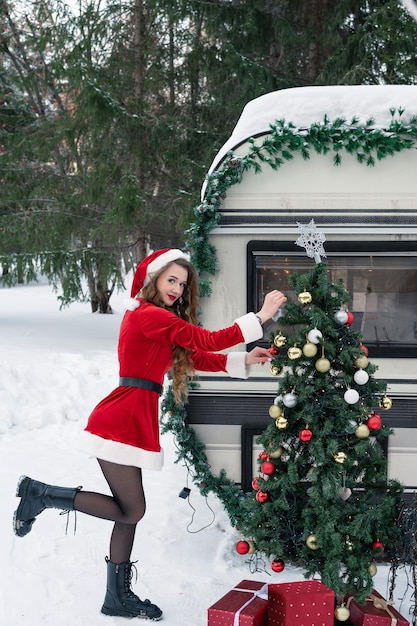 Junge Frau im Weihnachtsmannkostüm schmückt den Weihnachtsbaum auf dem Wintercampingplatz und bereitet sich darauf vor