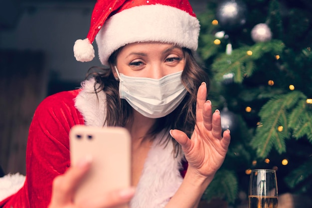 Junge Frau im Weihnachtsmannkostüm sitzt vor einem Smartphone und winkt mit den Händen. Weihnachtsgrüße im Internet. Neujahr im Quarantänekonzept.