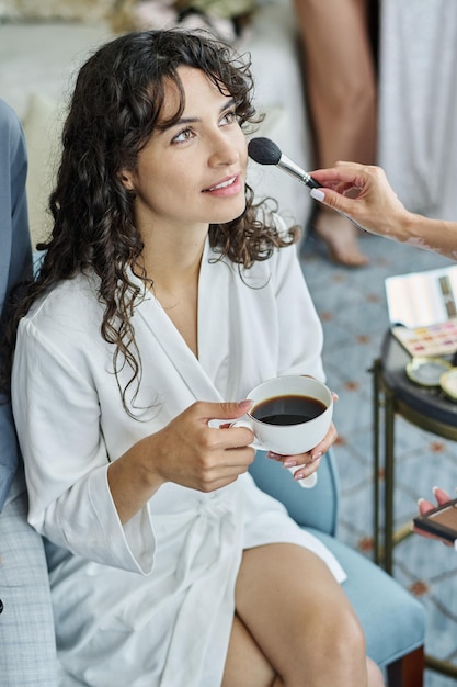 Junge Frau im weißen Bademantel, die während des Make-up-Verfahrens Espresso trinkt