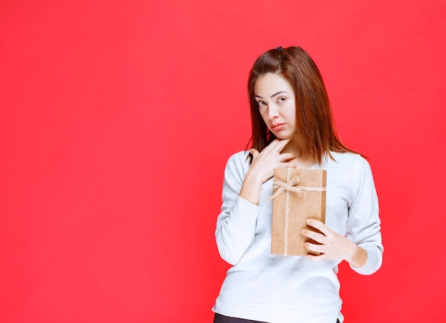 Junge Frau im weißen Hemd, die eine Geschenkbox aus Karton hält und verwirrt und nachdenklich aussieht