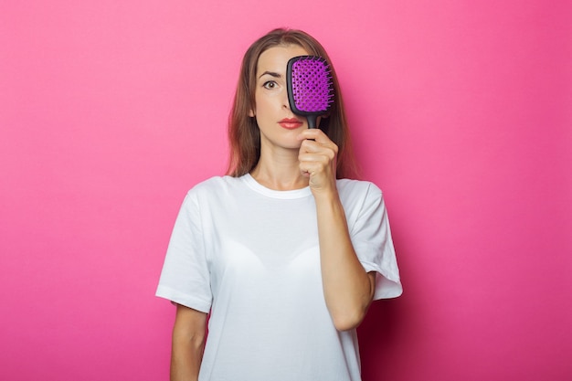 Junge Frau im weißen T-Shirt bedeckt ihr Gesicht mit Kamm auf Rosa.