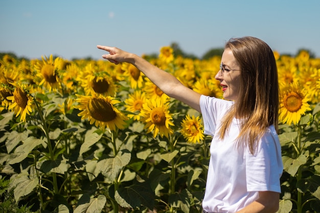 Junge Frau im weißen T-Shirt und in den Gläsern zeigt mit einem Finger auf einem Sonnenblumenfeld