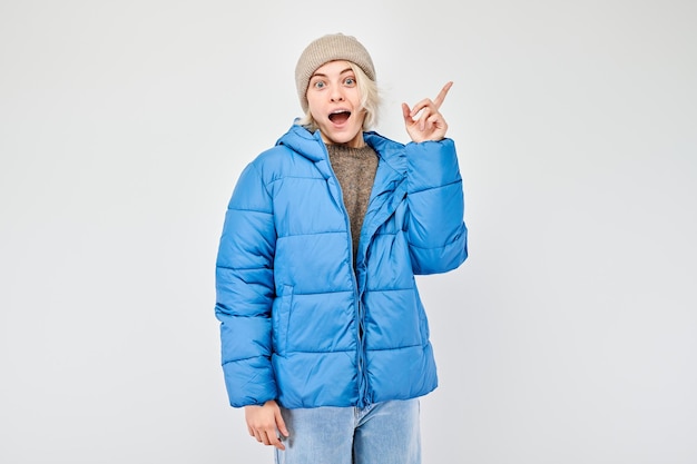 Junge Frau in blauer Winterjacke, die mit dem Finger zur Seite zeigt und einen leeren Raum für ein Produkt oder einen Text zeigt, der auf einem weißen Studio-Hintergrund isoliert ist