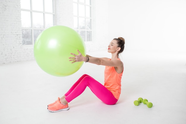 Junge Frau in bunter Sportkleidung, die sich mit Fitnessball in der weißen Turnhalle ausdehnt