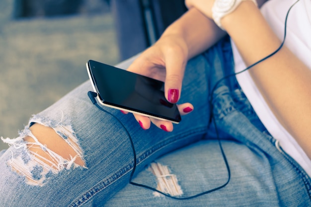 Junge Frau in den Jeans, die auf der Bank sitzen und schließen die Kopfhörer an ihren Handy an