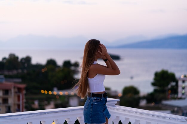 Junge Frau in den Jeansshorts, die auf dem Balkon stehen und den Meerblick und den schönen Sonnenuntergang betrachten. Urlaub auf tropischer Insel. Luxuskonzept.