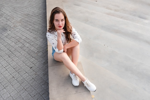 Junge Frau in Denim-Shorts, die am Wasser sitzt. Schönes Mädchen posiert auf einem Hintergrund der Stadt an einem sonnigen Tag. Mädchen in Freizeitkleidung.