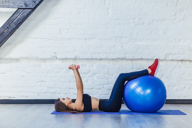 Junge Frau in der Fitnesskleidung, die mit Fitnessball und Hanteln, lokalisiert auf weißem Raum ausübt.