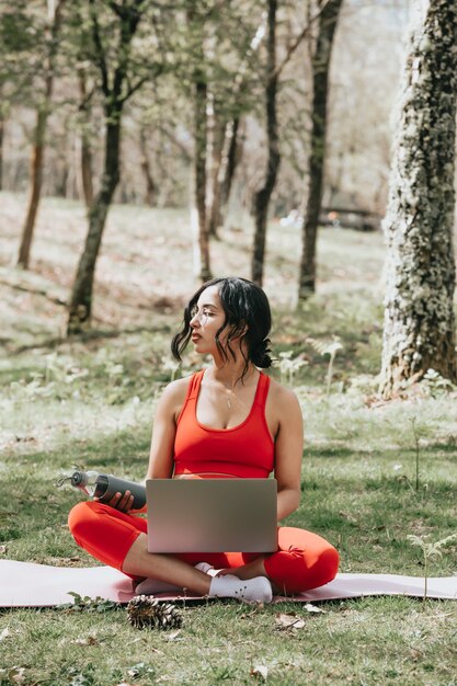 Junge Frau in der Fitnesskleidung, die sich auf Laptop entspannt, der eine Flasche Wasser am Park über einer Yogamatte ergreift