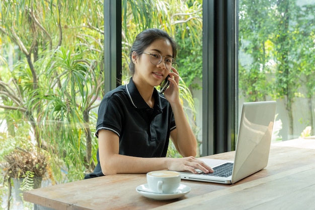 Junge Frau in der Kaffeestube unter Verwendung eines Laptops und Unterhaltung an einem Handy.