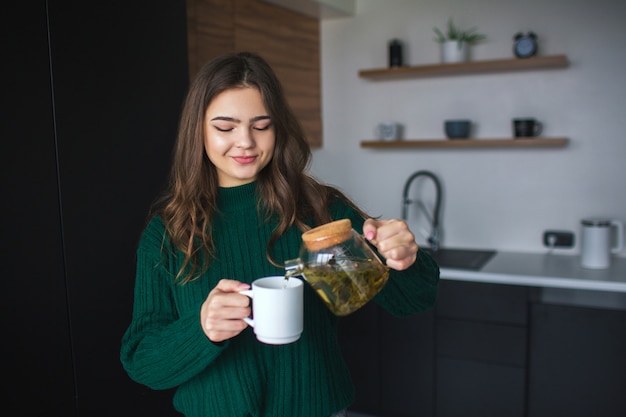 Junge Frau in der Küche während der Quarantäne. Mädchen, das etwas Tee von der Teekanne in die weiße Tasse gießt. Schöne Frau im grünen Pulloverlächeln. Allein in der Küche.