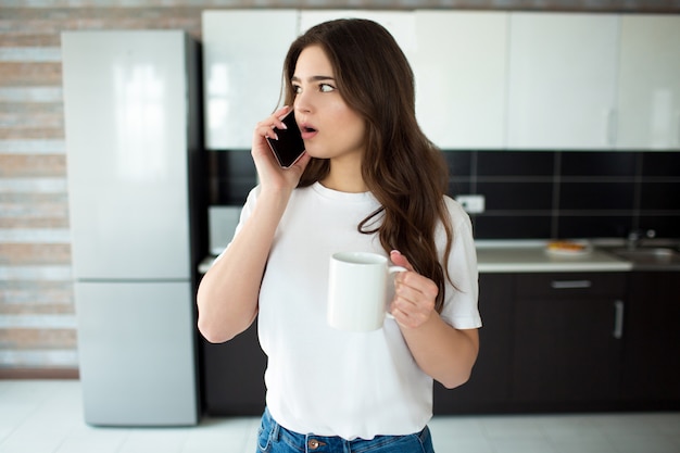 Junge Frau in der Küche. Weibliches Modell, das mit Staunen am Telefon spricht und nach links schaut. Weiße Tasse in der Hand halten. Tageslicht in der Küche.
