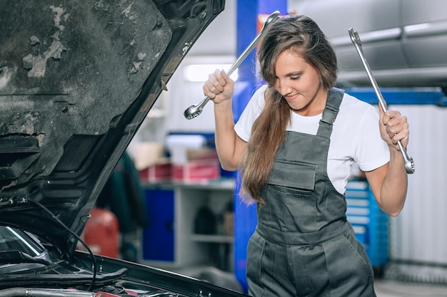 Junge Frau in der Nähe der offenen Motorhaube eines schwarzen Autos in der Garage. Das Mädchen trägt einen schwarzen Overall und ein weißes T-Shirt, sie hält die Schraubenschlüssel hoch und senkt den Blick.