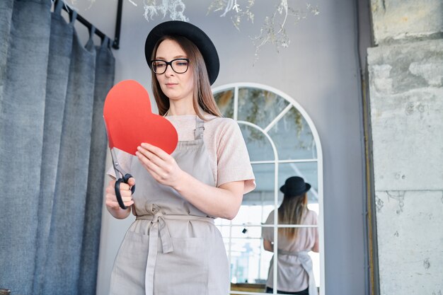 Junge Frau in der Schürze und im Hut, die großes rotes Papierherz und Schere halten, während sie für Valentinstag vorbereiten