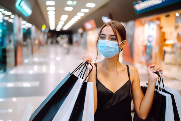 Junge Frau in der sterilen medizinischen Schutzmaske auf ihrem Gesicht mit Einkaufstüten im Einkaufszentrum.