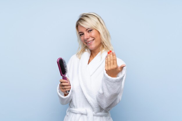 Junge Frau in einem Bademantel mit dem Haarkamm, der einlädt, mit der Hand zu kommen. Schön, dass Sie gekommen sind