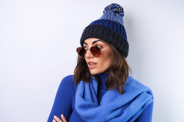 Junge Frau in einem blauen Golf-Turtleneck-Hut und Schal mit Sonnenbrille auf weißem Hintergrund