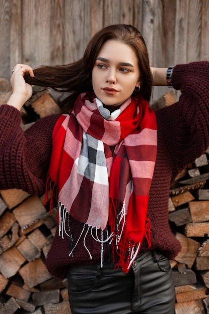 junge Frau in einem gestrickten Pullover mit einem karierten Schal in Lederhosen, der nahe einem alten Schuppen aufwirft