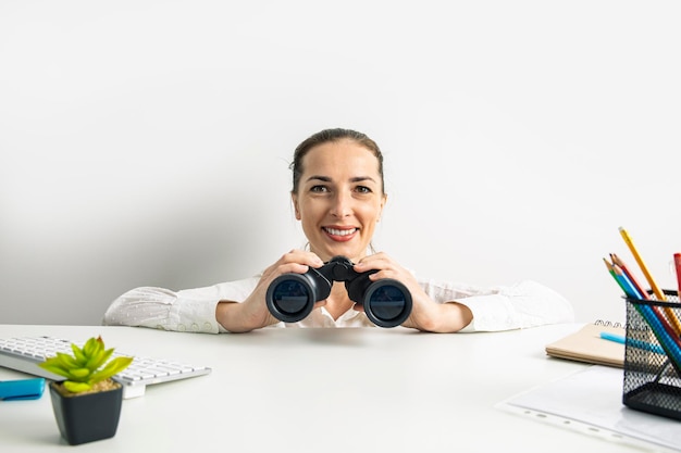 Junge frau in einem hemd mit fernglas, die hinter dem arbeitsplatz hervorschaut