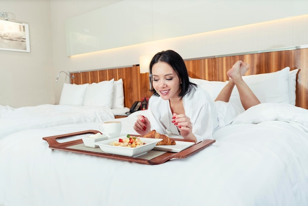 junge Frau in einem Hotel im Bademantel, die auf dem Bett liegt und mit Kaffee und Croissants frühstückt