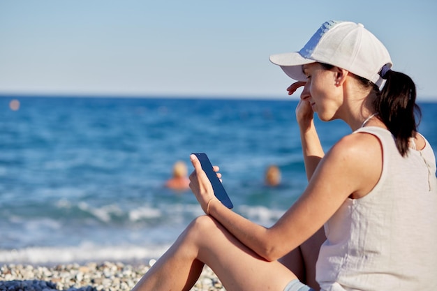 Junge Frau in einem Hut mit einem Telefon am Strand in der Nähe des Meeres Das Konzept der freiberuflichen Online-Online-Konferenz oder des Online-Studiums Hochwertiges Foto