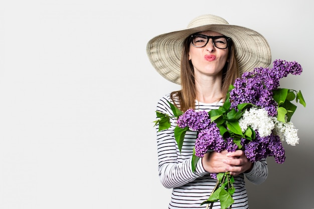 Junge Frau in einem Hut und in den Brillengrimassen, die einen Blumenstrauß auf einem hellen Hintergrund halten.