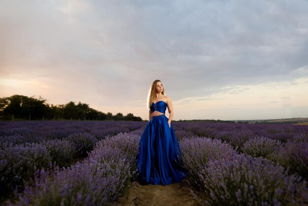 Junge Frau in einem luxuriösen blauen Kleid, das auf einem Lavendelfeld vor dem Hintergrund des Himmels steht.