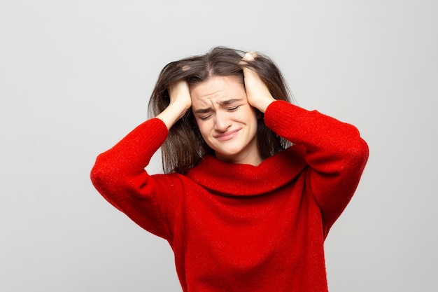 Junge Frau in einem roten Pullover mit starken Kopfschmerzen hält ihren Kopf isoliert auf einem weißgrauen Hintergrund