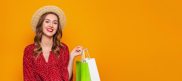 Junge Frau in einem roten Sommerkleid mit Strohhut hält eine Einkaufstüte isoliert auf orangefarbenem Hintergrund