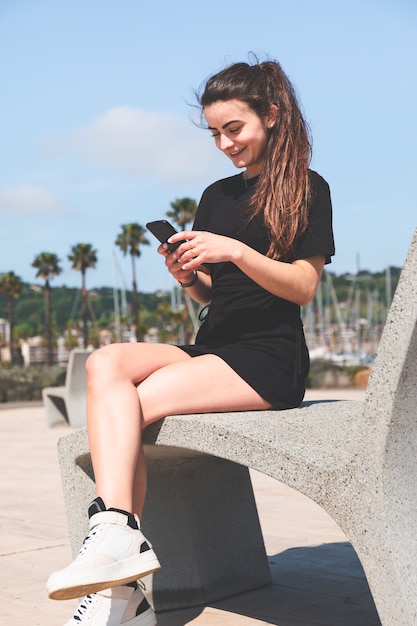 Junge Frau in einem schwarzen Kleid auf einer Bank mit einem Handy