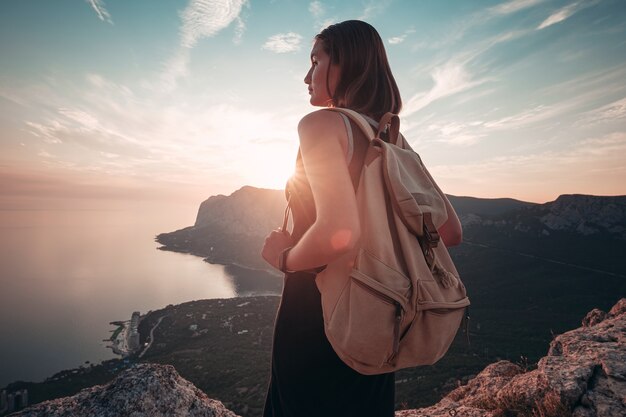 Junge Frau in einem Sportkleid mit Rucksack genießt den Blick auf die Berge und das Meer bei Sonnenuntergang. Reise- und aktives Lifestyle-Konzept. Glamping