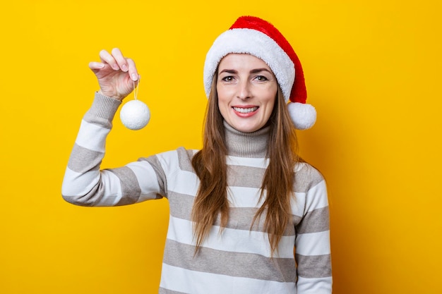 Junge Frau in einem Weihnachtsmann-Hut hält einen dekorativen Ball auf gelbem Hintergrund