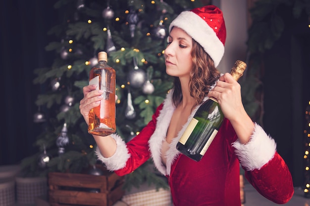 Junge Frau in einem Weihnachtsmann-Kostüm hält eine Flasche Champagner und Whisky. Wählen Sie ein alkoholisches Getränk für den Urlaub.