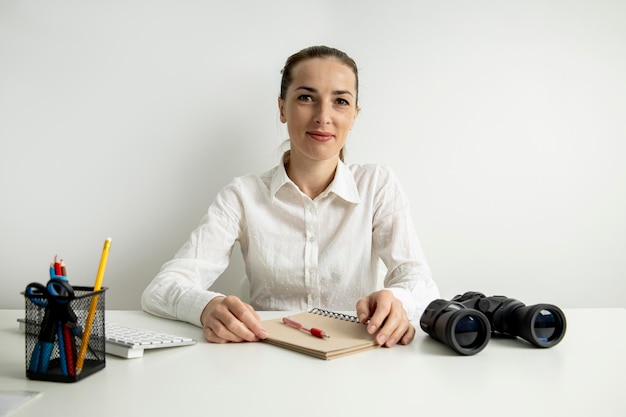 Junge Frau in einem weißen Hemd mit Fernglas, die im Büro sitzt