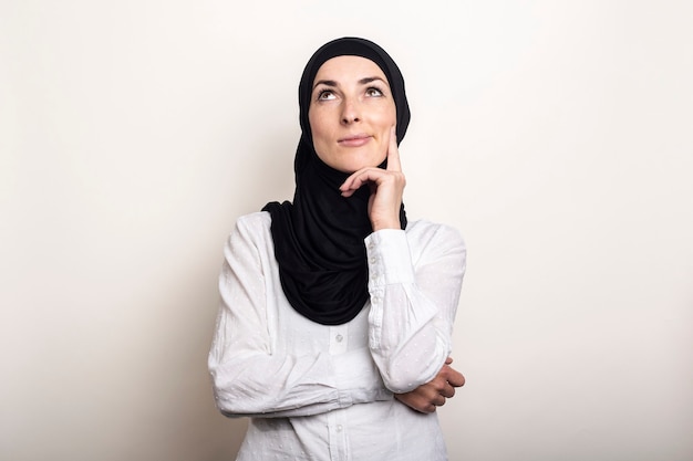 Junge Frau in einem weißen Hemd und Hijab hält ihre Hand an ihr Kinn und schaut mit einem nachdenklichen Gesicht auf