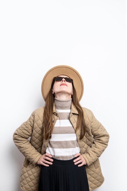 Junge Frau in einer Jacke mit Sonnenbrille in einem Hut vor einer weißen Wand