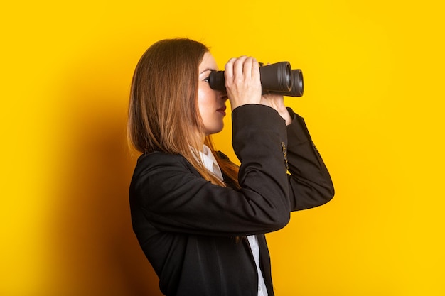 Junge Frau in einer Jacke schaut durch ein Fernglas auf gelbem Hintergrund