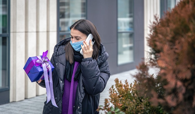 Junge Frau in einer Schutzmaske mit einem Geschenk in den Händen beim Telefonieren