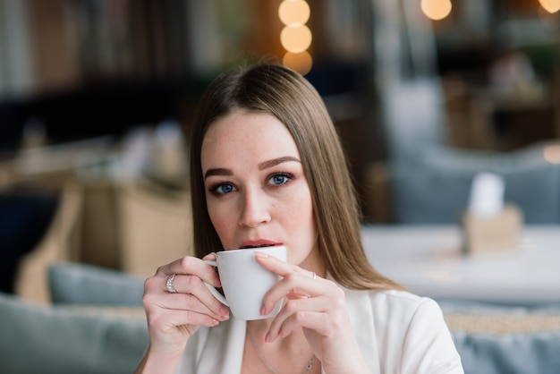 Junge Frau in einer weißen Jacke, die im Café mit Papieren arbeitet, Kaffee trinkend