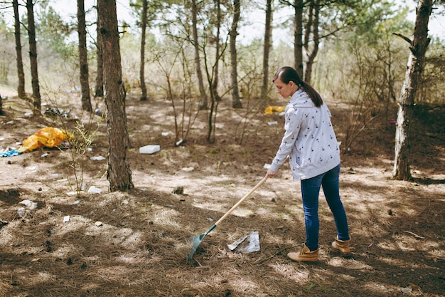 Junge Frau in Freizeitkleidung, die Müll mit Rechen für die Müllabfuhr in übersäten Parks oder Wäldern säubert. Problem der Umweltverschmutzung